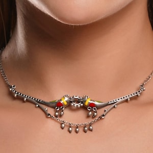 K&ouml;nnte beinhalten: Eine silberne Halskette mit zwei vogelartigen Anh&auml;ngern mit roten, gelben und gr&uuml;nen Akzenten. Die Halskette hat eine filigrane Kette mit kleinen, h&auml;ngenden Perlen. Das Design ist symmetrisch und liegt auf dem Hals einer Person.