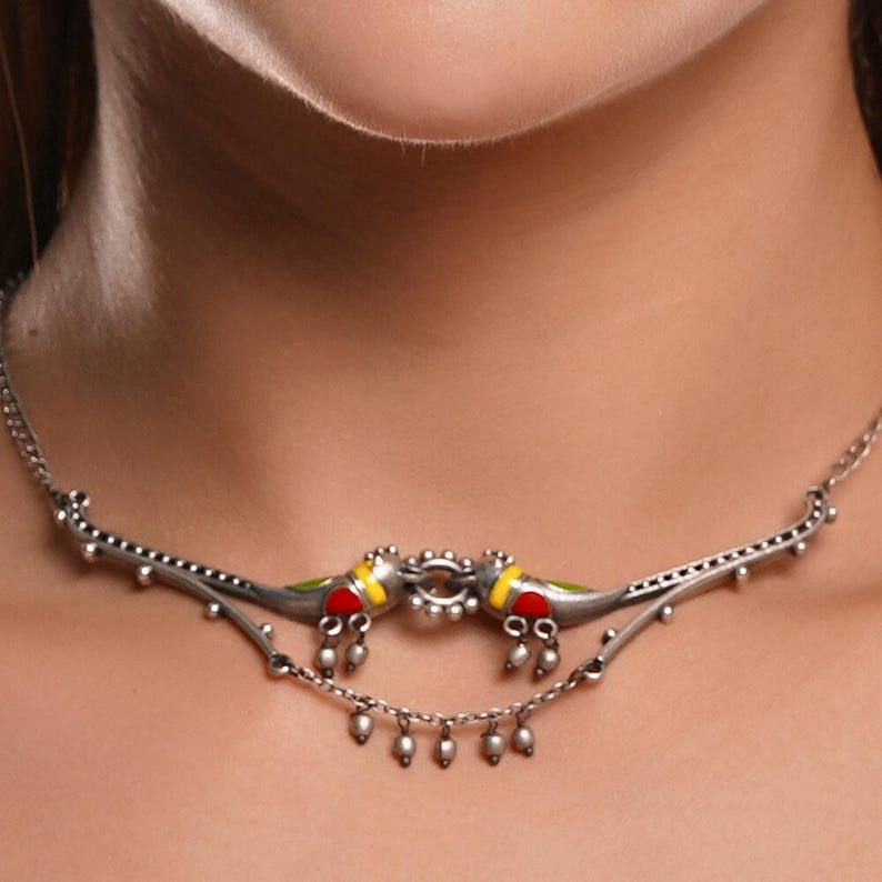 K&ouml;nnte beinhalten: Eine silberne Halskette mit zwei vogelartigen Anh&auml;ngern mit roten, gelben und gr&uuml;nen Akzenten. Die Halskette hat eine filigrane Kette mit kleinen, h&auml;ngenden Perlen. Das Design ist symmetrisch und liegt auf dem Hals einer Person.