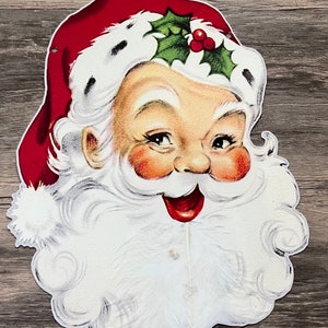 May include: A vintage paper cutout of Santa Claus's face with a red hat, white beard, and a green holly sprig with red berries.