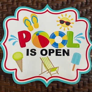 May include: A colorful sign with the text "Pool is Open" in black letters. The sign features a pool float, ice cream cone, beach chair, and flip flops. The sign is decorated with blue, red, and white accents.