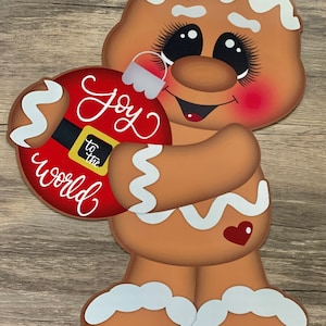 May include: A gingerbread man with a red and white ornament that says "Joy to the world". The gingerbread man is smiling and has rosy cheeks.