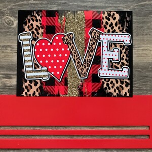 May include: A red wooden sign with the word "LOVE" in white letters with a red heart and leopard print accents. The sign is on a red wooden stand.