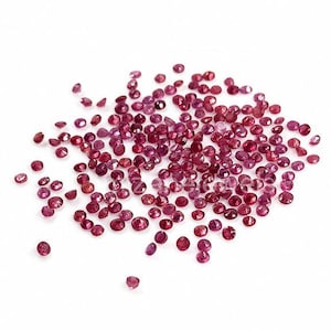 Op de afbeelding: Een verspreiding van kleine, gefacetteerde, dieprode edelstenen tegen een witte achtergrond. De edelstenen zijn gegroepeerd, met enkele verder verspreid. De afbeelding is goed belicht, wat de glans van de stenen benadrukt. Het logo "Mizane Jewels" staat rechtsonder.