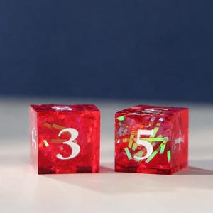 May include: Two translucent red six-sided dice made of resin, each containing embedded iridescent foil flakes. The die on the left displays the number three in white, while the die on the right displays the number five in white on its front face.