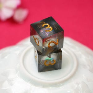 May include: Two stacked translucent dark gray resin dice with gold-colored numbers. Each die contains a spherical core filled with iridescent, multi-colored glitter particles. The dice are square-shaped with rounded edges and clear, polished surfaces.