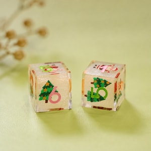 May include: Two translucent, glittery, cube-shaped dice featuring festive holiday graphics. Each die displays a green Christmas tree illustration alongside a large, bold number in pink or green ink. The dice have smooth, rounded edges and a clear finish.