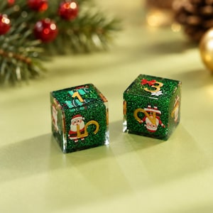 May include: Two square dice made of green glitter resin. Each die features gold-colored numbers and small illustrations of Santa Claus. One die displays the number 1, while the other shows the number 3. The dice have a smooth, glossy finish.