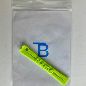 Pode incluir: Uma etiqueta de plástico verde-limão com um pequeno orifício circular numa extremidade e a palavra Name em relevo ao longo do centro. A etiqueta está posicionada na diagonal sobre uma inserção de papel branco dentro de um saco plástico.