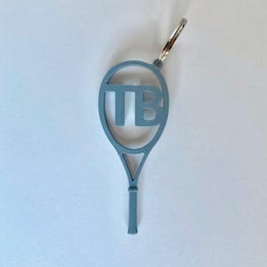 Puede incluir: Un llavero de raqueta de tenis azul claro con las letras "TB" en el centro. El llavero es de metal y tiene un llavero plateado en la parte superior. El mango de la raqueta está en la parte inferior.