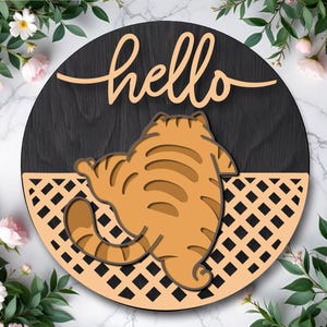 Puede incluir: Letrero redondo de madera con un gato naranja y marrón y la palabra "hello" en cursiva. El gato está acostado sobre un fondo negro con un diseño de celosía. El letrero está rodeado de flores rosas y vegetación.