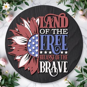Op de afbeelding: Een ronde, zwarte houten plaat met een patriottistisch zonnebloemontwerp. De bloemblaadjes zijn rood en wit, met een blauw centrum versierd met witte sterren. De plaat leest "Land of the Free Because of the Brave."