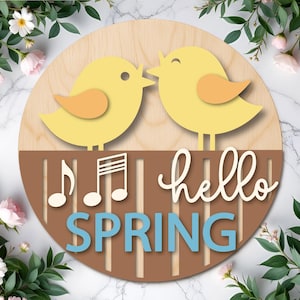 Op de afbeelding: Een ronde houten plank met twee gele vogels die elkaar kussen. Het bord zegt "hello SPRING" in wit en blauw, met muzieknoten. Het ontwerp is geplaatst tegen een achtergrond van groen en roze bloemen. Het bord is gemaakt van hout.