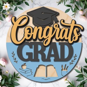 以下が含まれることがあります： 「Congrats GRAD」の文字が金と黒で書かれた丸い卒業祝いの木製サイン。卒業帽子、リンゴ、本、鉛筆が描かれています。サインは緑とピンクの花で囲まれています。