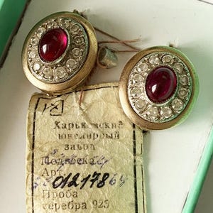 May include: Vintage gold-toned earrings with a central oval ruby surrounded by a halo of clear gemstones. The earrings are round and set in a gold-colored metal. A tag with Cyrillic text is visible.