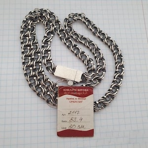 May include: A silver chain necklace with a complex interlocking link pattern and a rectangular clasp. A small red and white tag is attached, featuring the text "ЮВЕЛІРНІ ВИРОБИ", "ФОП АНЬЩУК А.Й.", "Україна, м. Вінниця", "СРІБЛО 925", "Арт. 2117", "Вага 53.4", and "60 см".