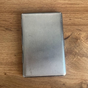 May include: A rectangular, silver cigarette case with a textured, cross-hatched pattern. The closed case sits on a wooden surface. The case has a slight sheen, reflecting the light.
