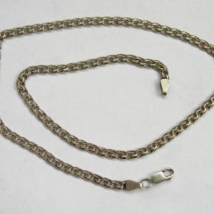 May include: A silver-toned metal chain necklace featuring a continuous round link design. The chain is arranged in a loose, circular loop and includes a small metal lobster claw clasp at one end and a flat, tapered metal end piece at the other.