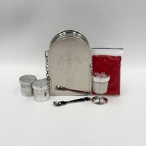 May include: A silver-toned metal liturgical set featuring an arched case with an engraved crucifix, two small lidded containers, a chalice with a cross on the lid, two small spoons, and a clear plastic bag containing a red powder substance.