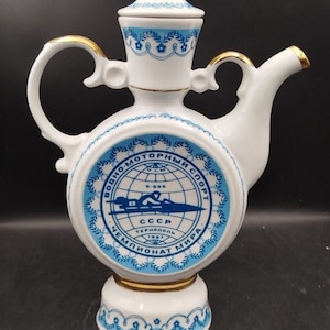 May include: A white ceramic teapot with blue floral patterns and gold accents. The teapot features a round body with a blue emblem and text in Cyrillic. The lid and handle are also decorated with gold trim.