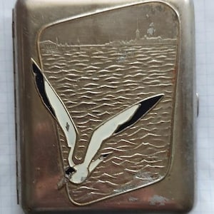 May include: A silver-coloured metal cigarette case with an embossed design. The case depicts a seagull with black and white wings, set against a water scene and a distant cityscape. The case is rectangular with rounded corners.