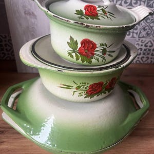 Puede incluir: Juego de tres piezas de esmalte en tonos verdes y blancos, con un diseño floral de rosas rojas. El conjunto incluye un cuenco grande con asas, una olla mediana y una olla más pequeña con tapa. Las piezas tienen un aspecto vintage.