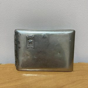 May include: A rectangular, silver-toned metal cigarette case. The case has a hinged lid and a decorative emblem with the word "HIMIS" on the front. The case is closed and sits on a wooden surface.