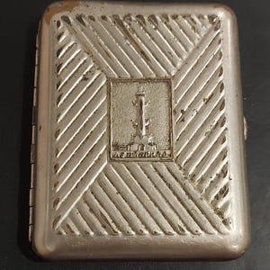 May include: A rectangular silver-toned metal cigarette case featuring a diagonal striped pattern and a central embossed square emblem depicting a monument with the Cyrillic text LENINGRAD inscribed at the base of the architectural structure.