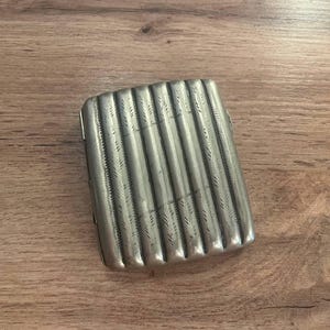 Könnte beinhalten: Eine silberne, rechteckige Zigarettenetui mit geripptem Design. Das Etui hat eine strukturierte Oberfläche mit vertikalen Linien und aufwendigen Details. Das Etui ist geschlossen und liegt auf einer Holzoberfläche.