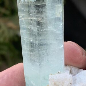 May include: A translucent, light blue aquamarine crystal with a textured, crystalline structure. The crystal is held against a blurred background, showcasing its natural form and color. The base is a white, rough mineral.