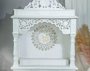 May include: A white marble temple with detailed carvings. The structure has a decorative arch with a gold Om symbol surrounded by sparkling embellishments. A small, white, flower-shaped dish is in front of the temple.