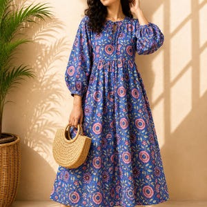May include: A blue midi dress featuring a repeating floral pattern in shades of pink and white, long puff sleeves with elastic cuffs, a round neckline with a tie closure, and a gathered waistline, paired with a woven straw handbag and sandals.