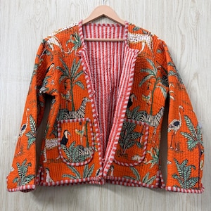 Op de afbeelding: Een gewatteerd jack in een levendige oranje kleur met een herhalend patroon van zebra's, toekans en tropisch gebladerte. Het kledingstuk heeft lange mouwen, twee opgestikte voorzakken en een rood-wit gestreepte bies langs de randen.