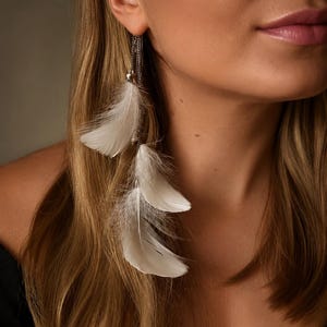 Pode incluir: Um close-up de um brinco pendurado com três penas brancas suspensas por uma corrente delicada. O brinco está preso à orelha de uma mulher. As penas têm uma aparência macia e texturizada, e a corrente tem um brilho metálico.