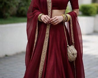 May include: A deep burgundy Indian outfit, comprising a crop top, long skirt, and sheer scarf. The ensemble is embellished with gold detailing and accessorized with gold bangles and a small, decorative purse.