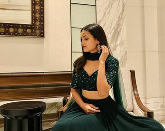 May include: A dark green two-piece formal outfit. The top is a cropped wrap style with short sleeves and small white embellishments. The skirt is a long, flowing design. The outfit is worn with matching accessories.
