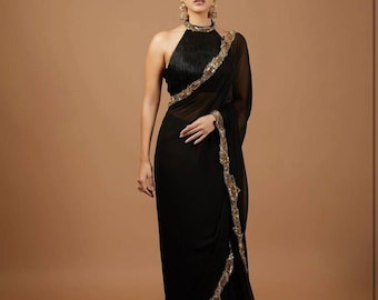 Puede incluir: Un sari negro con cuello halter y adornos dorados. El sari presenta un diseño fluido con un borde decorativo. La modelo lleva pendientes y un anillo dorados.