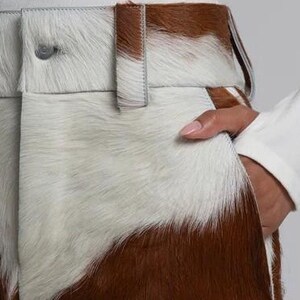 May include: Close-up of brown and white cowhide trousers. The trousers have a button closure and belt loops. The white and brown pattern is visible on the front. A hand is in the pocket. The trousers are part of a clothing collection.