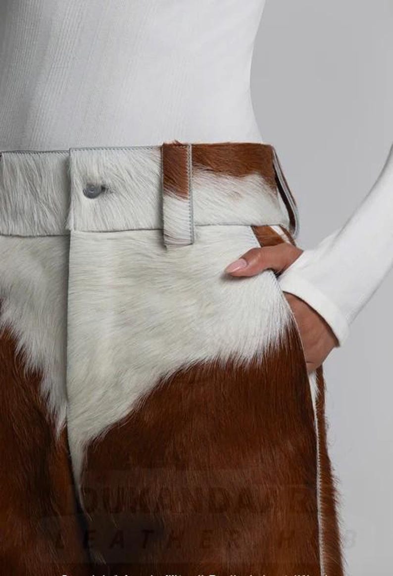 May include: Close-up of brown and white cowhide trousers. The trousers have a button closure and belt loops. The white and brown pattern is visible on the front. A hand is in the pocket. The trousers are part of a clothing collection.