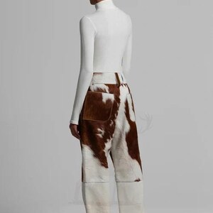 May include: A person wearing a white turtleneck top and cowhide trousers. The trousers feature a brown and white pattern, with a brown pocket on the back. The trousers have a wide-leg design.
