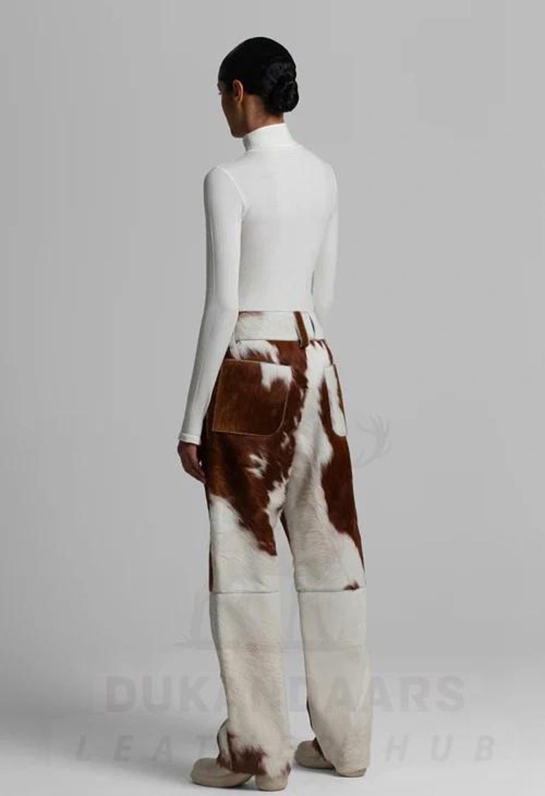 May include: A person wearing a white turtleneck top and cowhide trousers. The trousers feature a brown and white pattern, with a brown pocket on the back. The trousers have a wide-leg design.