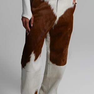 May include: Cowhide trousers in brown and white, featuring a distinctive pattern. The trousers have a matching belt and are made from a soft, textured material. Beige shoes complete the look.