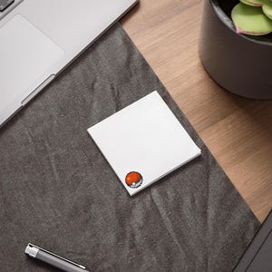 May include: A stack of white square sticky notes featuring a pixelated red and white design. The notes are on a dark grey surface, with a laptop and pen visible. A potted plant is also in the background, on a wooden desk.