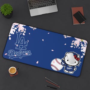 Puede incluir: Una alfombrilla de escritorio rectangular con fondo azul oscuro y detalles de flores de cerezo blancas. El diseño incluye el logotipo blanco de los LA Dodgers, una pelota de béisbol y Hello Kitty con una camiseta de los Dodgers y un ratón blanco.