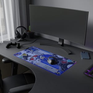 Puede incluir: Alfombrilla de ratón rectangular para juegos con el personaje Sonic the Hedgehog. La alfombrilla tiene un borde azul con un diseño colorido, un ratón de ordenador y un teclado en un escritorio oscuro.