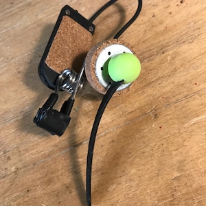 May include: A mechanical device featuring a circular cork base with a bright lime green sphere, a metal spring, a black rectangular component with a cork inlay, and a flexible black cord, all connected by thin metal wires and black tubing.