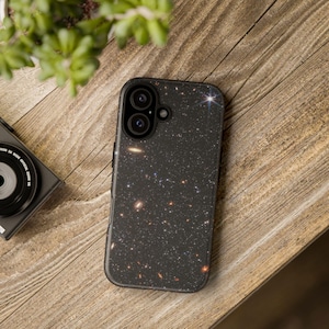 May include: Black smartphone case featuring a starry galaxy design. The case has a dual-lens camera cutout and a sleek, modern appearance. The case is set on a wooden surface, with a camera and plant in the background.