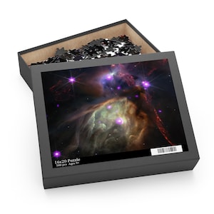 Peut inclure: Une boîte de puzzle de 40 x 50 cm avec une image vibrante de nébuleuse spatiale. La boîte est ouverte, révélant des pièces de puzzle. L'image présente des couleurs violettes, vertes et brunes, avec des points brillants en forme d'étoiles. La boîte noire porte l'inscription "16x20 Puzzle 500 pcs Ages 9+".