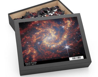 May include: A black puzzle box with a lid open, revealing a partially assembled puzzle. The puzzle image features a vibrant depiction of a galaxy with swirling orange, red, and blue hues, and bright white stars. The box reads "16x20 Puzzle" and "500 pcs Ages 9+".