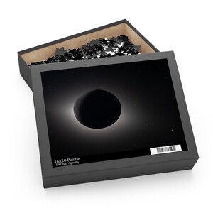 May include: A black puzzle box with a partially open lid, revealing a partially assembled puzzle. The box features a black and white image of a solar eclipse. Text on the box reads "16x20 Puzzle 500 pcs Ages 9+".