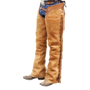 May include: Tan suede leather chaps with fringe detailing. The chaps are worn over blue jeans and have a fitted design with a decorative panel on the thigh. These chaps are designed for riding and western wear, and are made of suede leather.